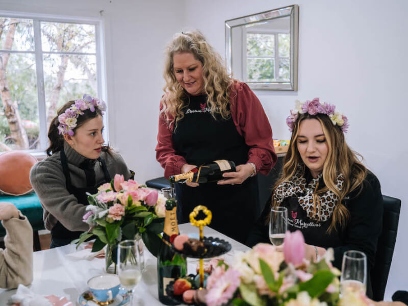 Three women make flower crowns and pour champagne at a table with fresh flowers